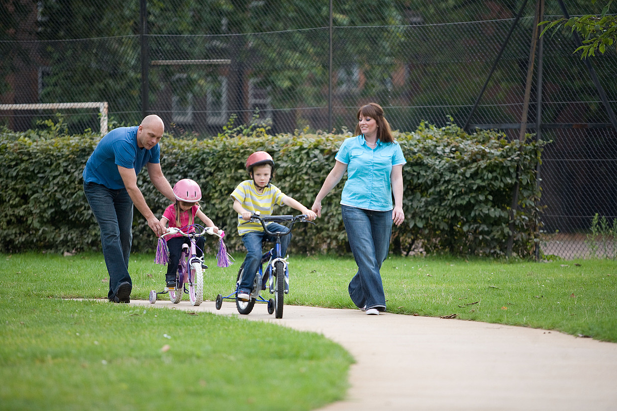 Unique ID: POSL1044 Caption: Outdoors in the park. A family going for a walk. Exercise. Two children riding bicycles. Wearing safety helmets. Cycling. Two adults. Parents and child. Active lifestyle. Fresh air. Restrictions: NHS Photo Library - for use in NHS, local authority Social Care services and Department of Health material only Copyright: ©Crown Copyright