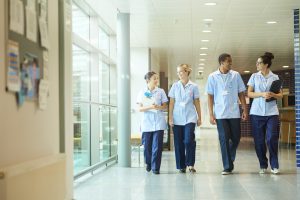 a group of four young trainee nurses including male and female nurses , walk towards camera down a hospital corridor . They are wearing uk nurse uniforms of trousers and tunics.