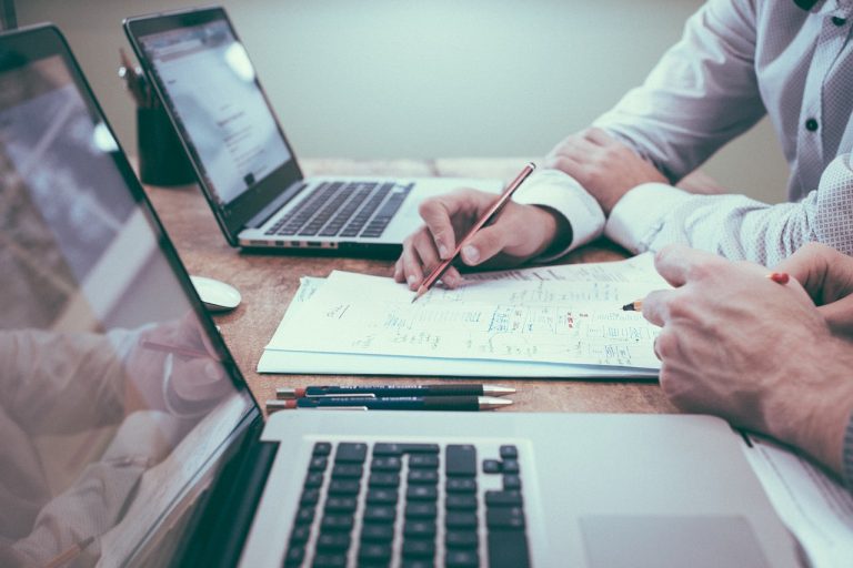 Two laptops on a desk with paper between them and two people's hands holding pens