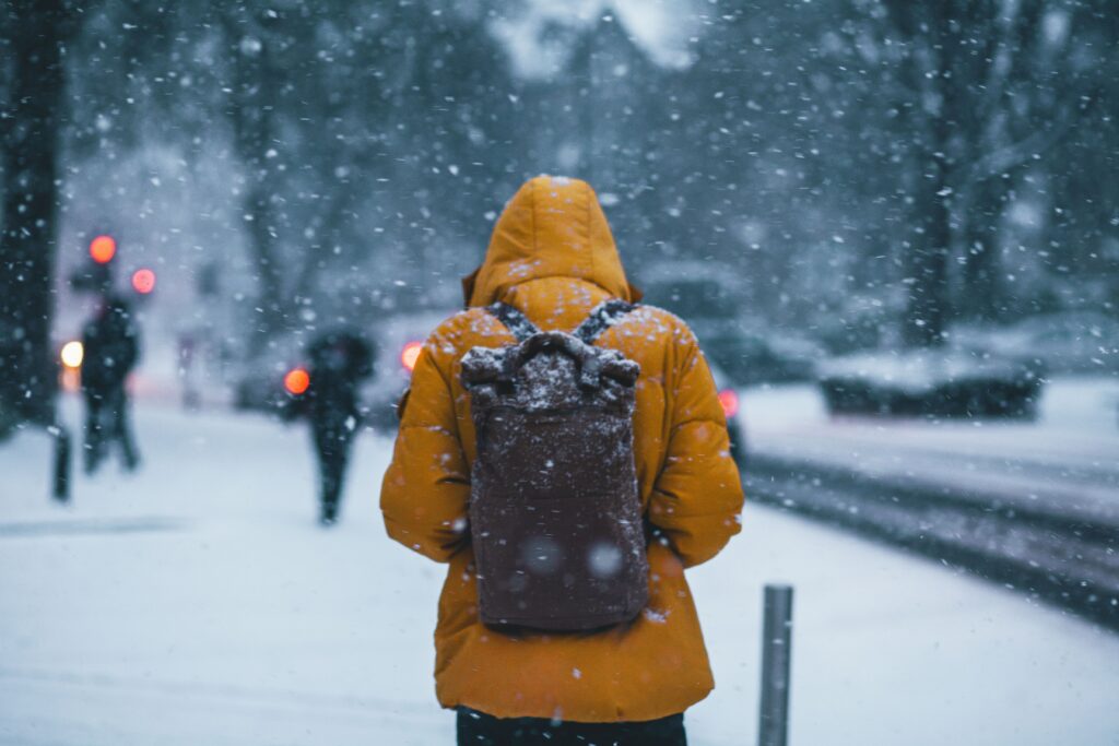 A person photographed from behind wearing a yellow winter coat, with the hood up and a brown backpack on their back. They are walking on a snow-covered street.
