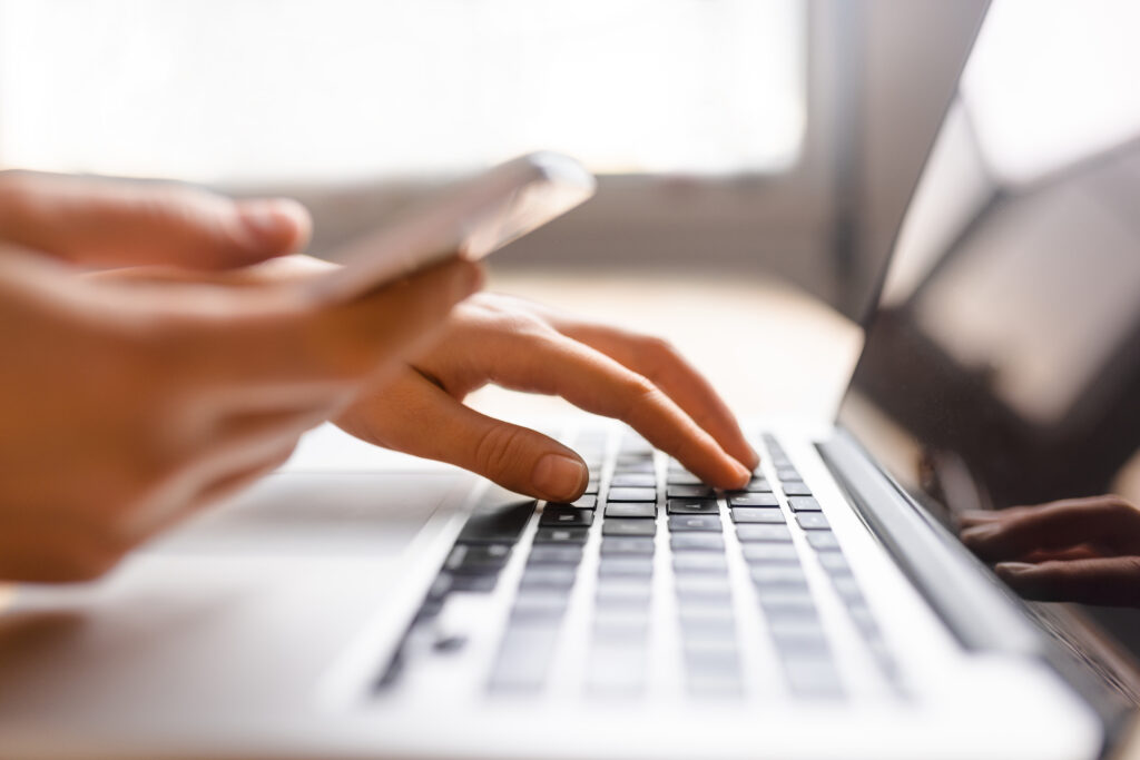 A pair of hands, one on a keyboard and one holding a phone in a white case. A laptop screen can be seen to the left of the image