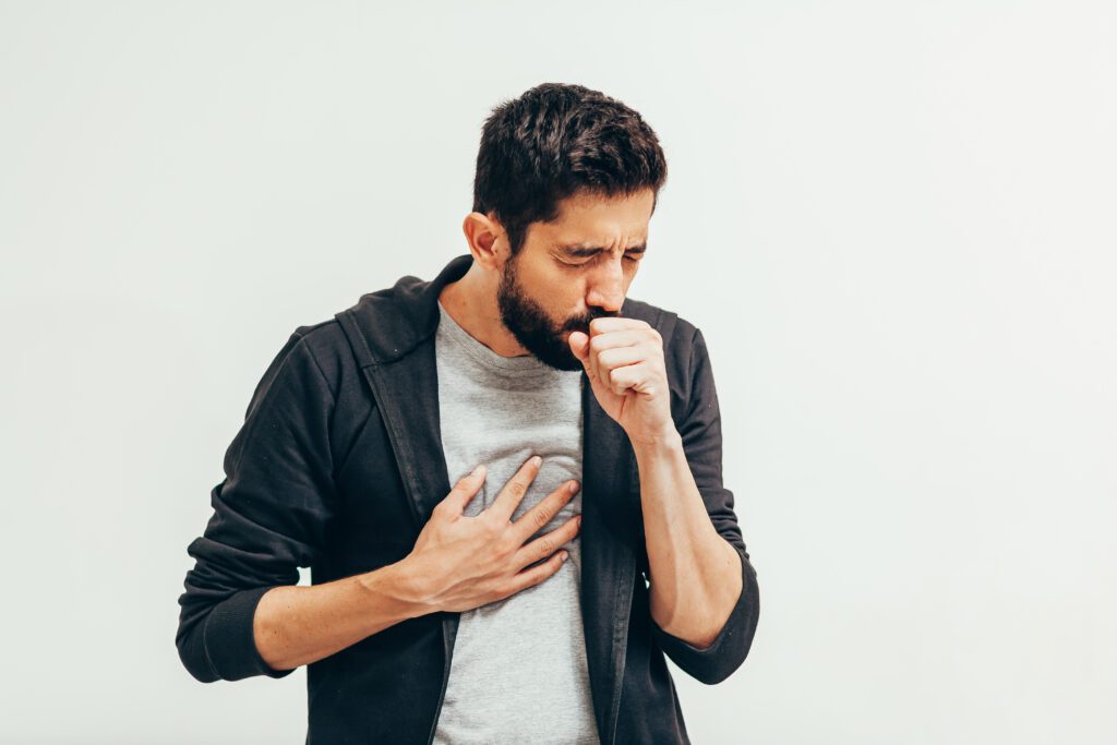 Man in black cardigan coughing with a hand over his mouth and the other hand on his chest.