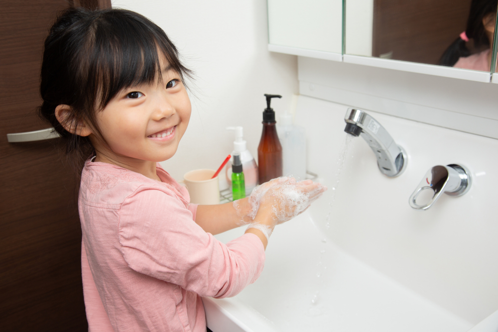 Young child standing at sink washing soap from her hands.