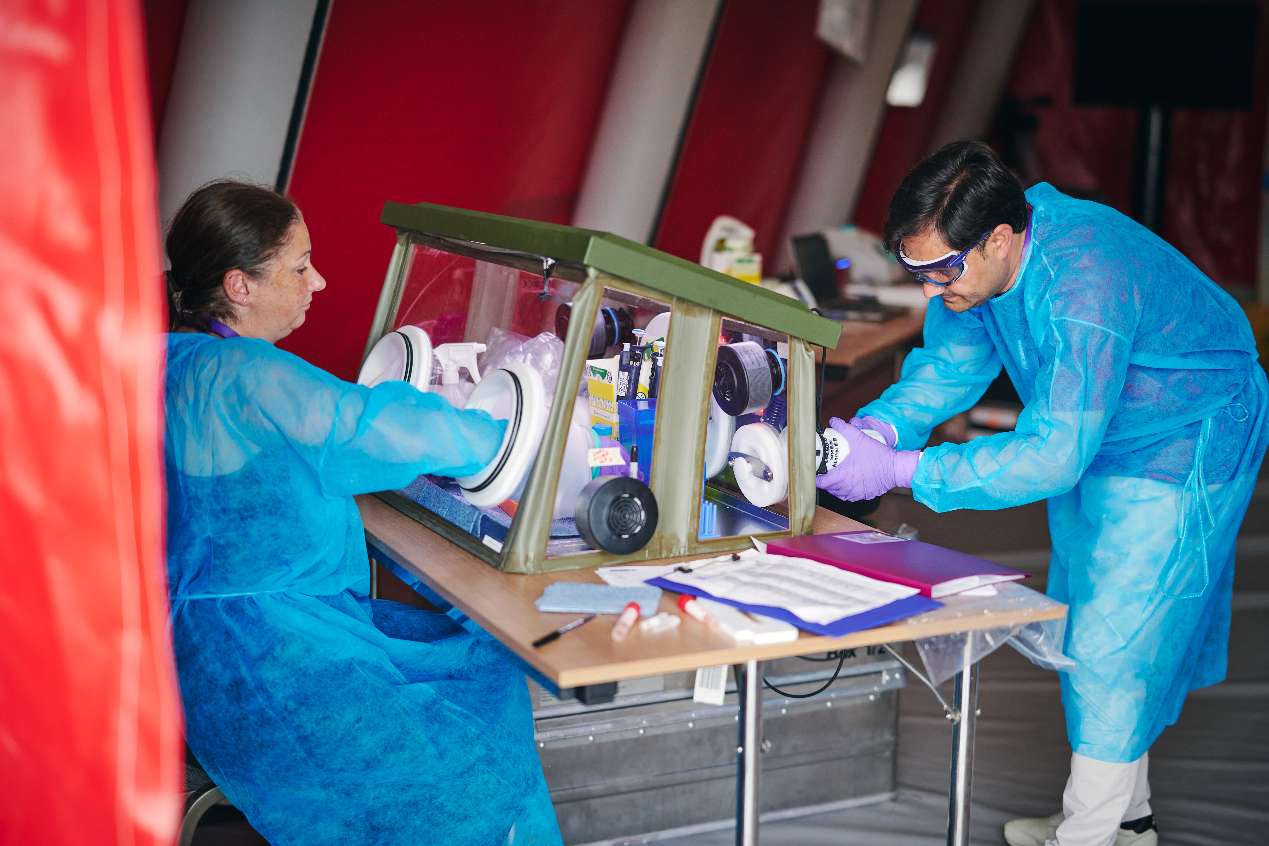 Two scientists, wearing protective clothing, using a foldable lab whilst in a tent.