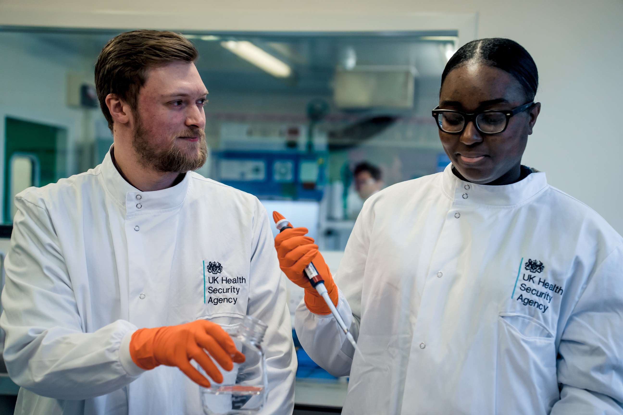 Picture of two UKHSA staff in a lab, wearing white lab coats and orange gloves