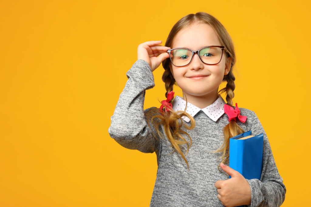 Child in glasses with plaits in hair holding a book and standing against a yellow background. 