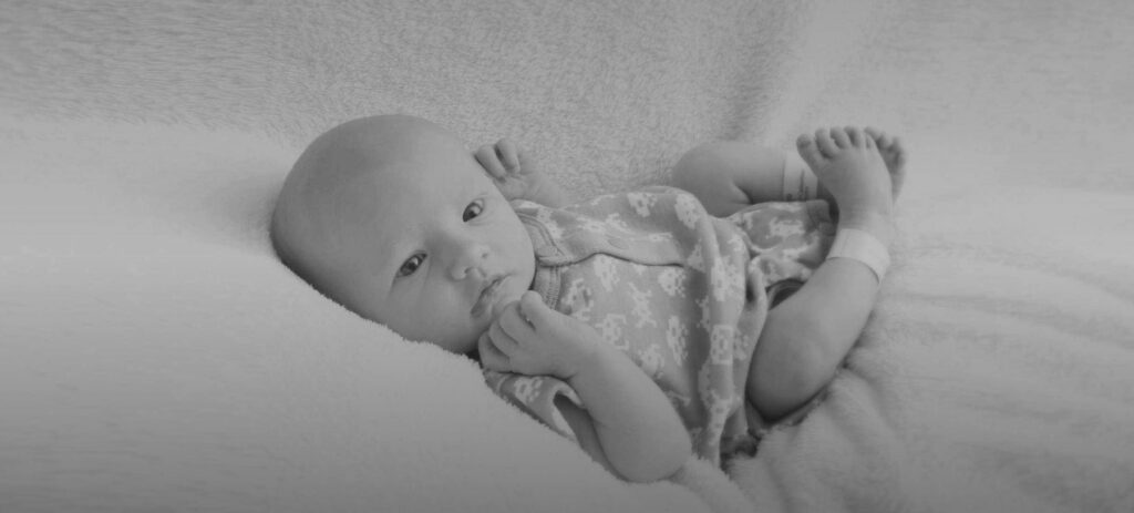 A grayscale image of a baby, Riley Hughes, wearing a singlet, looks towards the camera.