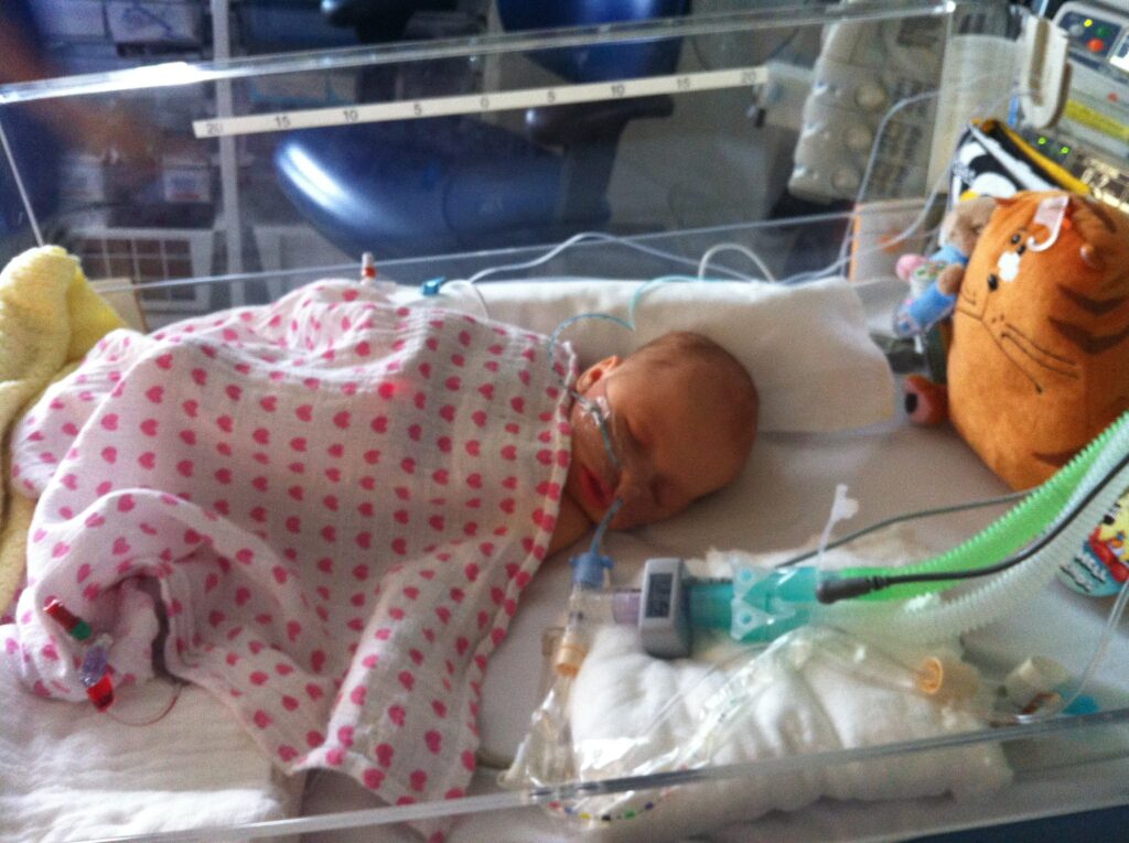 A three week old baby, Layla, intubated in a hospital bed, covered with a red spotted blanket.