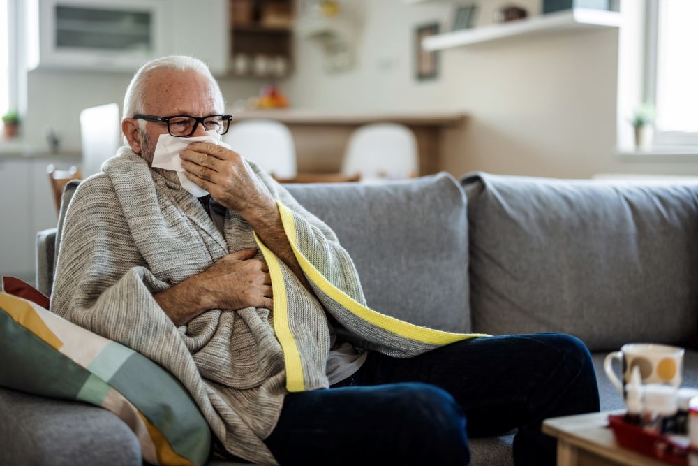 Elderly man catches a sneeze in a tissue, sitting on a grey sofa