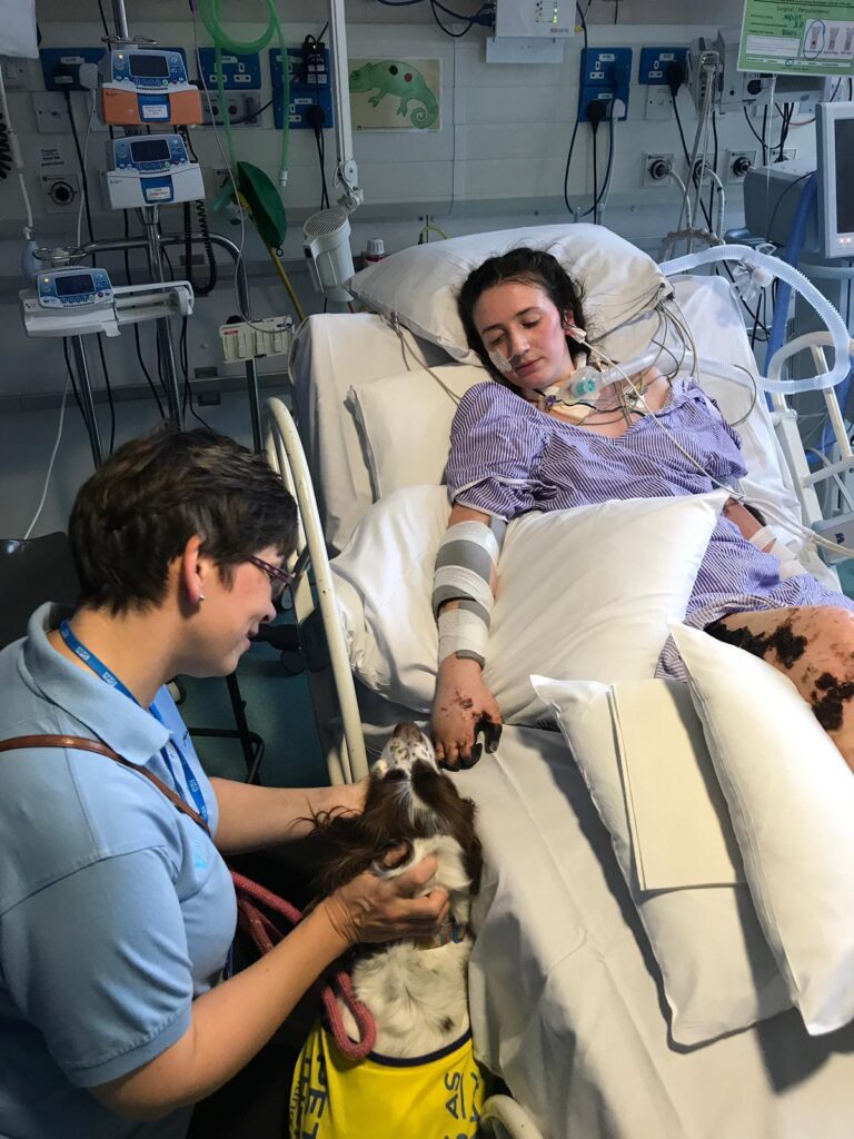 Becca Heritage, lying in her hospital bed, being visited by a dog and member of hospital staff. 

Becca's fingers, and patches on her legs, are covered in purpuric areas which look like large very dark areas of skin.