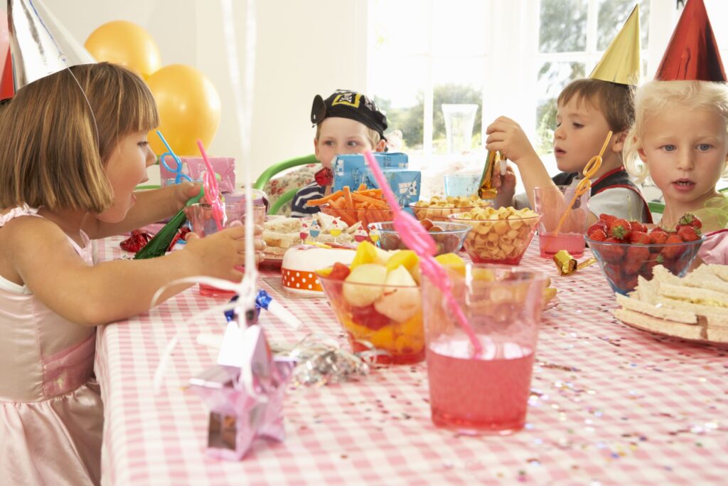Children sit around a table set for a birthday party, drinking juice and eating cake