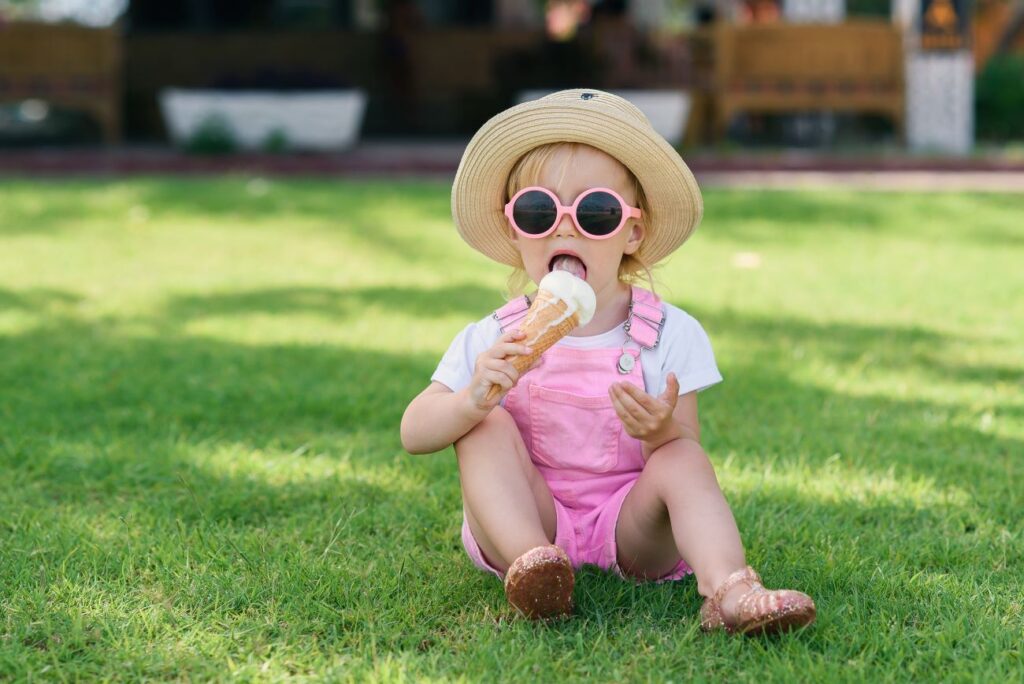 Little girl wearing hat, sunglasses and a pink romper suit, sits on grass eating an ice cream