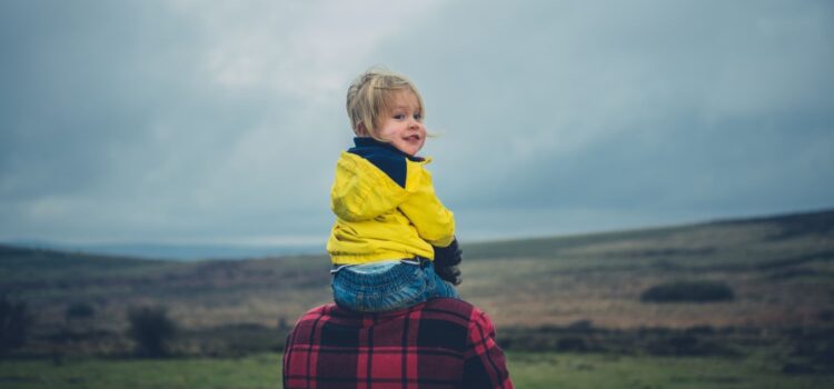 A toddler in a yellow mac sits on an adult's shoulders, against the backdrop of a cloudy sky