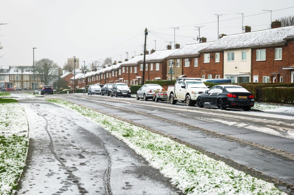 A residential street lined with brick houses on a snowy day. Light snow covers the roofs, pavements, and grassy areas, while the road is partially slushy. Several parked cars are lined up along the roadside, and the sky is overcast, giving the scene a cold, wintry atmosphere.