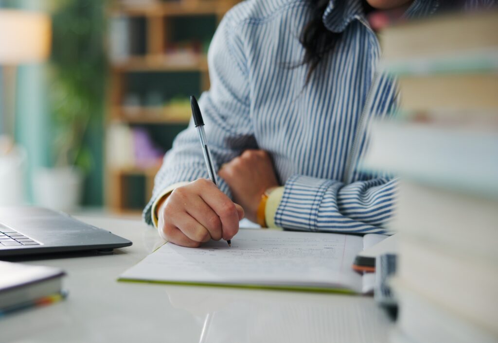 Student sitting, writing at a table, surrounded by books