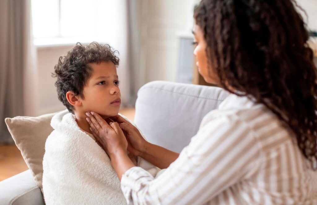 Person sitting on a sofa gently checks a child's throat while the child, wrapped in a white blanket, sits indoors near a bright window
